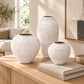 Three textured ceramic vases in matte off-white and light beige tones displayed on a wooden console table in a softly lit living room setting.
