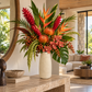 A vibrant tropical floral arrangement in a textured cream ceramic vase, displayed on a stone console table in a modern, sunlit living room.