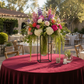 A large floral centerpiece arrangement in a clear glass vase with a black metal frame stand, placed on a round table covered with a deep burgundy tablecloth. The flowers include white, pink, purple, and peach blooms with green foliage.