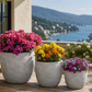 Three round gray concrete planters of different sizes holding green herbs, arranged on a wooden table in an outdoor patio setting with a scenic sea and mountains in the background.