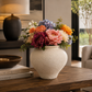 A textured cream-colored ceramic vase filled with a colorful assortment of artificial flowers, including pink, orange, purple, and white blooms with green foliage, displayed on a wooden table in a cozy living room setting.