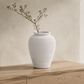 A textured white ceramic vase with a single delicate green branch, displayed on a light wood console table against a warm beige plaster wall.
