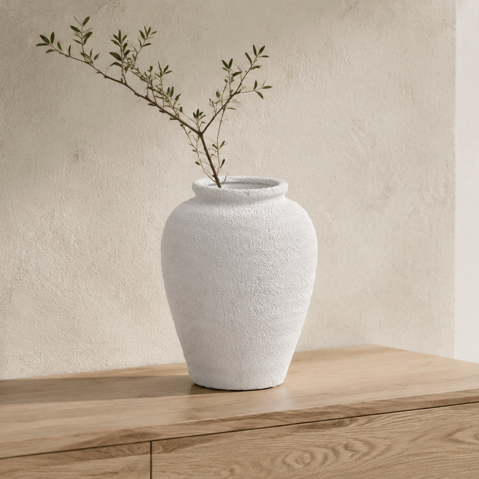 A textured white ceramic vase with a single delicate green branch, displayed on a light wood console table against a warm beige plaster wall.