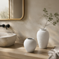Two textured white ceramic vases displayed on a light wood vanity next to a stone vessel sink, with a beige plaster wall and a folded linen towel in the background.