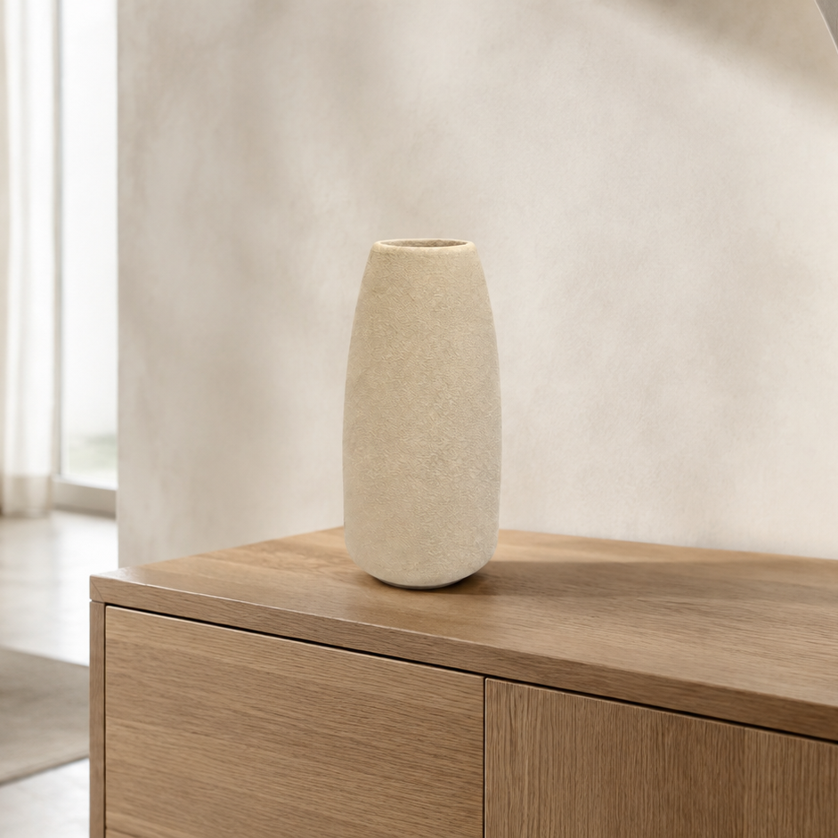 A tall, textured cream-colored ceramic vase sits on a light wood sideboard in a modern, minimalist room with warm neutral tones.