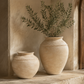 Two large beige terracotta-style decorative vases with rope detailing, one small and one tall, holding green olive branches, displayed in a rustic stone corridor.