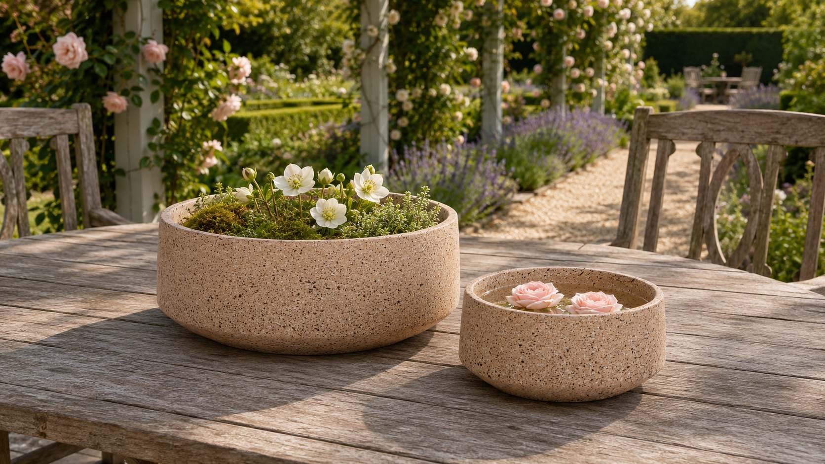 Two round terracotta-colored concrete planters sit on a weathered wooden table in a garden setting. The larger planter is filled with green foliage and small white flowers, while the smaller planter holds pink roses.