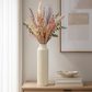 A tall cream-colored textured ceramic vase filled with a colorful arrangement of dried grasses, flowers, and greenery, styled on a light wood console table next to books and a small dish.