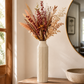 A tall cream-colored faceted ceramic vase filled with a dried floral arrangement in muted terracotta, beige, and peach tones, styled on a wooden console table in a warm, sunlit room next to a mirror and stone bowl.