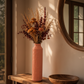 A tall, ribbed terracotta pink ceramic vase filled with dried autumn flowers, displayed on a rustic wooden console table in a warm, sunlit room.