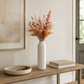 A tall, ribbed white ceramic vase filled with dried orange, pink, and brown botanical stems, styled on a wooden console table in a warm, neutral-toned room.