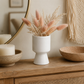 A white ceramic vase with a tiered base holding dried blush pink pampas grass, styled on a wooden console table with a candle, woven bowl, and macrame wall hanging.