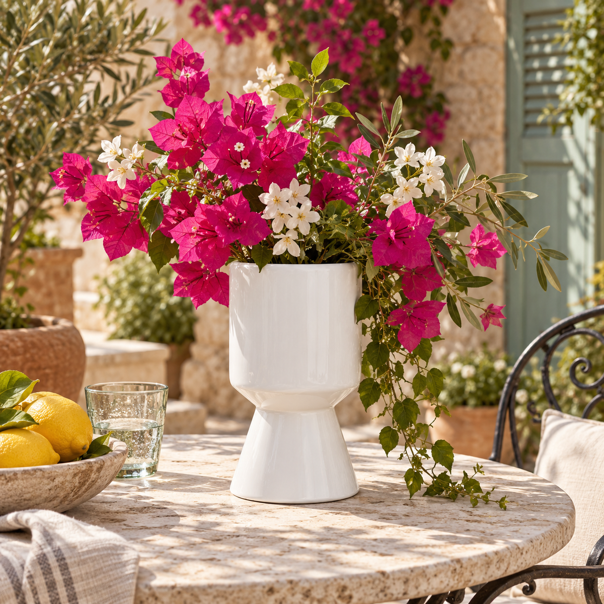 A white ceramic vase filled with bright pink and white flowers sits on a stone patio table outdoors, surrounded by greenery, lemons, and a glass of water.