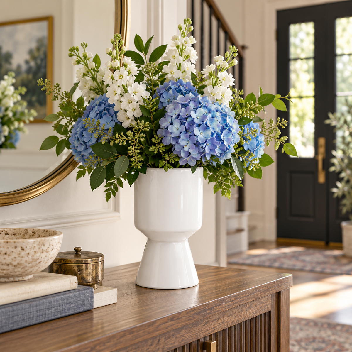 A white ceramic vase filled with a lush floral arrangement of blue hydrangeas, white flowers, and green foliage, displayed on a wooden console table in a bright, elegant entryway.