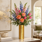 A vibrant floral arrangement featuring pink roses, tulips, blue delphiniums, and green foliage in a textured gold cylindrical vase, displayed on a marble-topped console table in a modern living room setting.