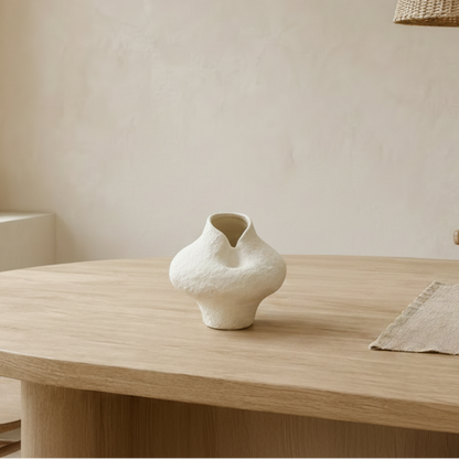 A small textured off-white ceramic vase with a bulbous shape sits on a light wood dining table in a minimalist room with beige walls, wooden chairs, and a woven pendant light.