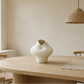 A textured off-white ceramic vase with a bulbous shape sits on a light wood dining table in a minimalist, warm-toned room with a woven pendant light and wooden chairs.