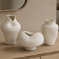 Three textured off-white ceramic vases displayed on a light wood vanity in a modern bathroom setting.