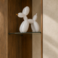 A white balloon dog sculpture displayed on a glass shelf within a wooden cabinet, next to a textured beige wall.