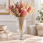 A clear ribbed glass pedestal vase filled with dried floral arrangements in pink, burgundy, cream, and muted green, displayed on a light stone table in a softly styled interior setting.