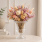 A clear glass urn-shaped vase filled with a dried floral arrangement in soft pastel tones, placed on a light stone table against a neutral indoor background.