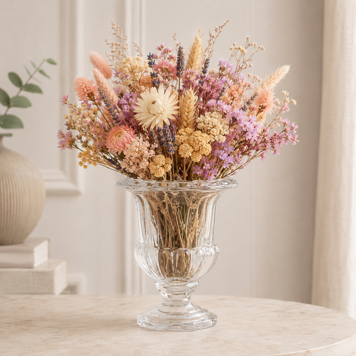 A clear glass urn-shaped vase filled with a dried floral arrangement in soft pastel tones, placed on a light stone table against a neutral indoor background.