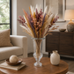 A dried flower arrangement in a clear glass pedestal vase, displayed on a round wooden coffee table in a modern living room setting.