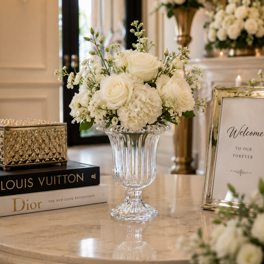 A white floral arrangement in a clear ribbed glass pedestal vase sits on a marble table in an elegant indoor setting. A framed 'Welcome to Our Forever' sign is beside it, with books including Louis Vuitton and Dior stacked underneath, and a decorative gold box on the left.