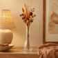 A clear glass vase filled with dried wheat, orange flowers, and burgundy blossoms, displayed on a wooden table in a warmly lit room next to a lamp and framed artwork.