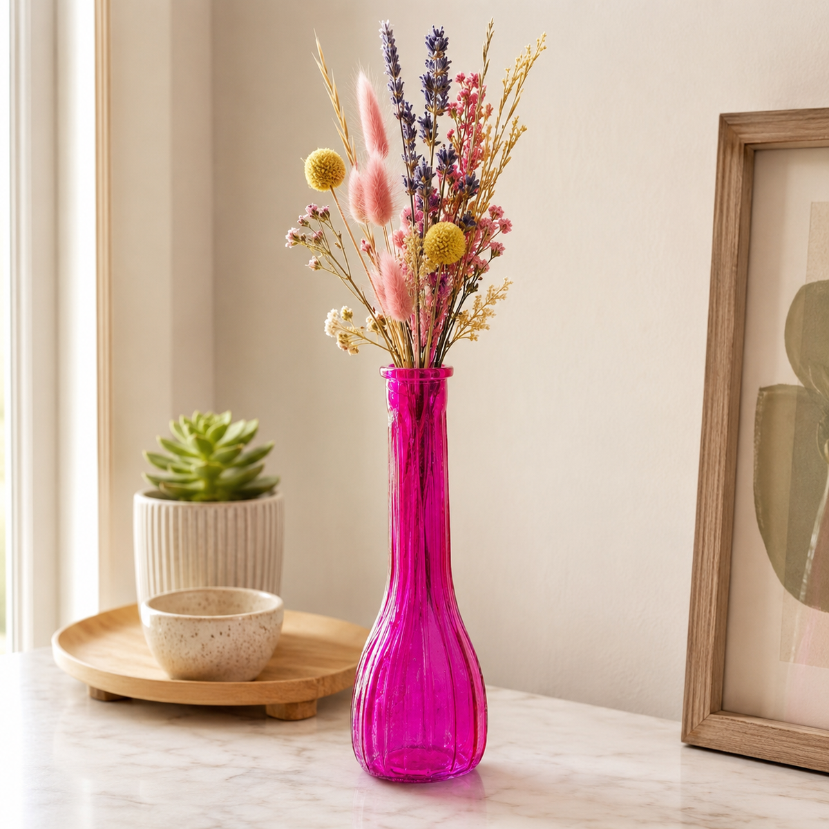 A bright magenta pink glass vase filled with dried flowers, set on a marble countertop in a styled home interior next to a small potted succulent and a framed print.