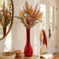 A tall, ribbed red glass vase filled with a dried floral arrangement in warm autumn tones, displayed on a wooden console table in a bright, modern entryway.