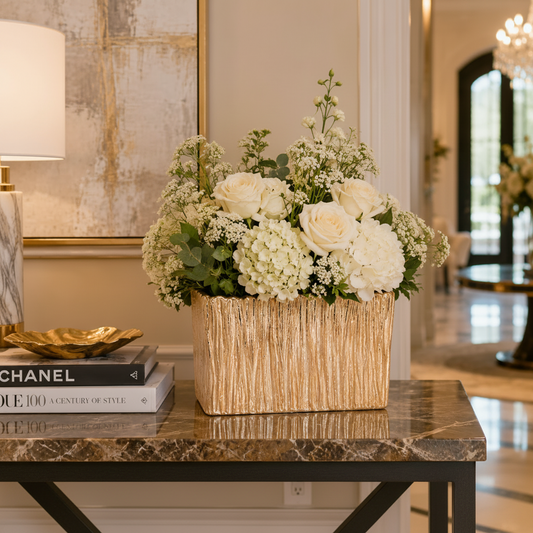 A luxury floral arrangement of white roses, hydrangeas, and eucalyptus in a textured gold rectangular vase, displayed on a marble console table in an elegant interior setting.