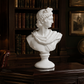 A white classical-style bust sculpture of Queen Elizabeth II, displayed on a dark wooden desk inside a traditional, dimly lit study with bookshelves, a lamp, and a globe in the background.