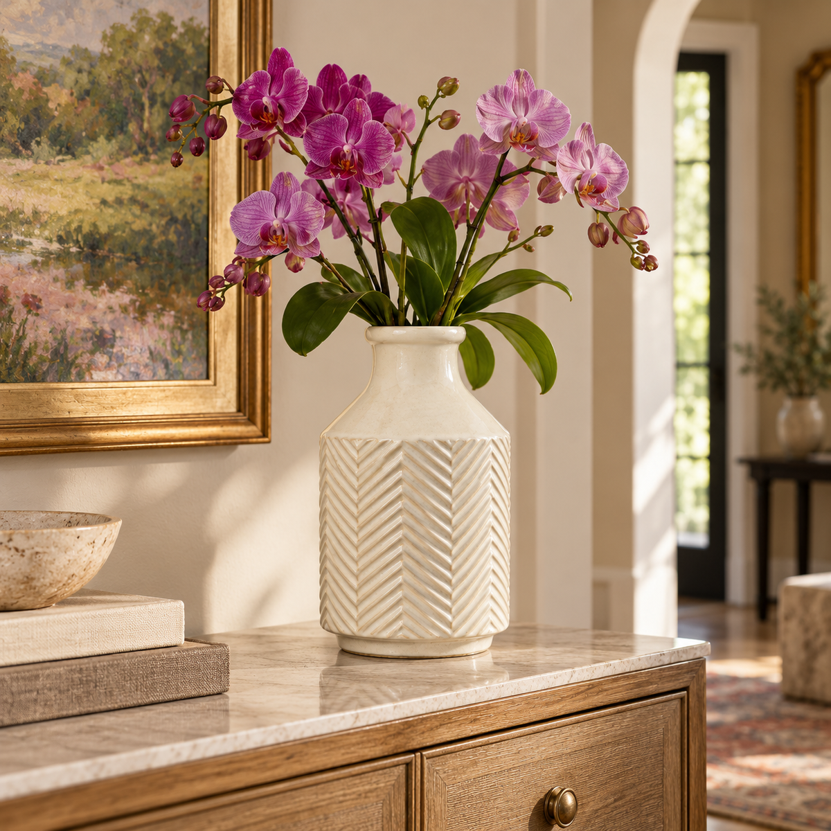 A white ceramic vase with a textured chevron pattern, filled with pink and purple orchids, displayed on a marble-topped console table in a warm, sunlit room.