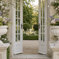 Two large white stone urn planters filled with blush pink and lavender flowers, placed on matching pedestals on either side of open white French doors in a garden setting.