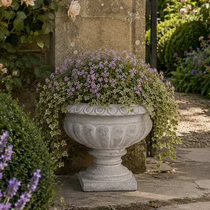 A large gray stone urn planter filled with trailing ivy and small purple flowers, placed outdoors against a stone pillar in a garden setting.