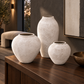 Three textured ceramic vases in cream and off-white tones displayed on a dark wood console table in a modern interior setting.