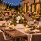 A beautifully set outdoor dining table at dusk with a large holiday centerpiece in a white ribbed vase, surrounded by lit candles, place settings, and festive greenery. A modern house with warm lights and decorated Christmas trees is visible in the background.