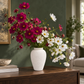 A white textured ceramic vase filled with a large arrangement of cosmos flowers, including deep pink, burgundy, and white blooms with yellow centers, displayed on a dark wood console table in a living room setting.