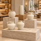 Five cream-colored ceramic vases in various shapes and sizes, displayed on matching stone pedestals in a modern, warm-toned living room setting.