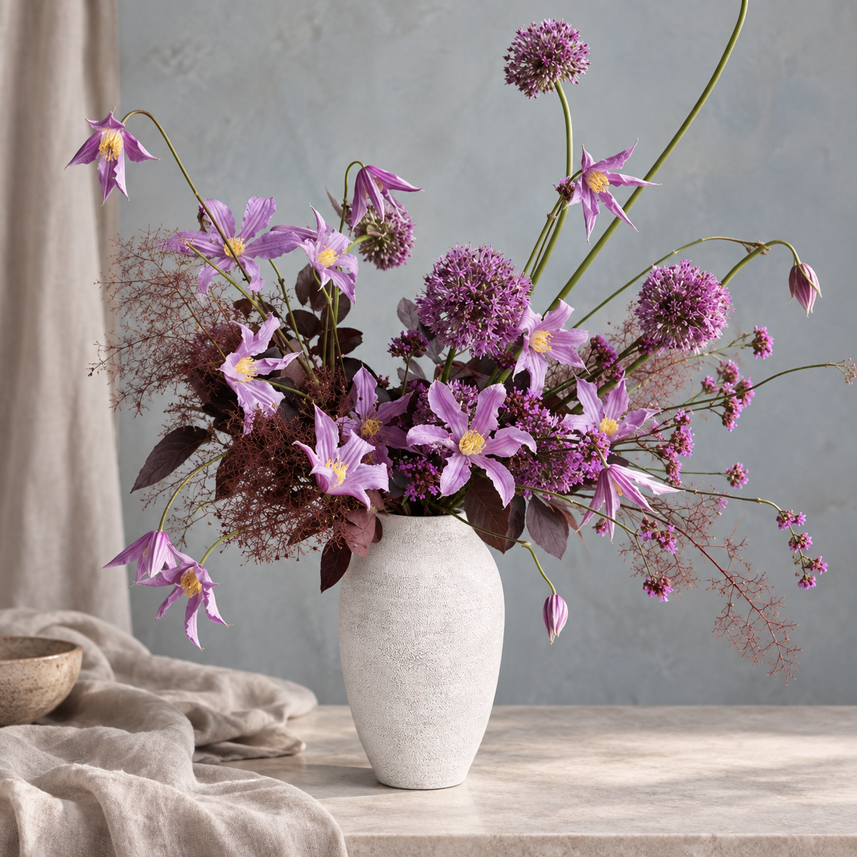 A dried floral arrangement in a textured off-white ceramic vase, featuring purple clematis flowers, allium-like spheres, and dusty burgundy filler stems, set on a stone tabletop with draped linen and a textured gray wall in the background.