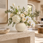 A large cream-colored ceramic vase filled with a lush floral arrangement of white orchids, cream roses, tulips, and hydrangeas, styled on a stone console table in a bright, modern living room.
