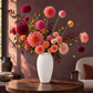 A vibrant floral arrangement of pink, red, and orange dahlias in a textured white ceramic vase, displayed on a dark wooden table in a warm, modern living room setting.