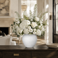 A large white ceramic vase filled with a lush white floral arrangement featuring hydrangeas, roses, ranunculus, and green foliage, displayed on a dark wood console table in a bright, elegant living room.