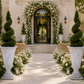 A beautifully styled wedding venue entrance with a stone pathway leading to an arched doorway decorated with white flowers and greenery. Large white urn planters with sculpted topiary trees flank the aisle, with wooden chairs arranged on both sides.