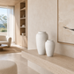 Two textured off-white ceramic vases displayed on a light stone console table in a modern, minimalist living room with warm beige walls and wood shelving.