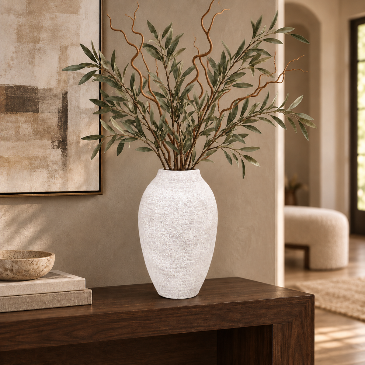 A textured white ceramic vase filled with green olive-leaf branches and tan decorative twigs, displayed on a dark wood console table in a warm, modern living room.